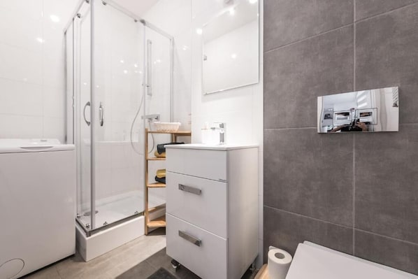 Un baño moderno con cabina de ducha de vidrio, tocador blanco con espejo y pared revestida de azulejos grises.