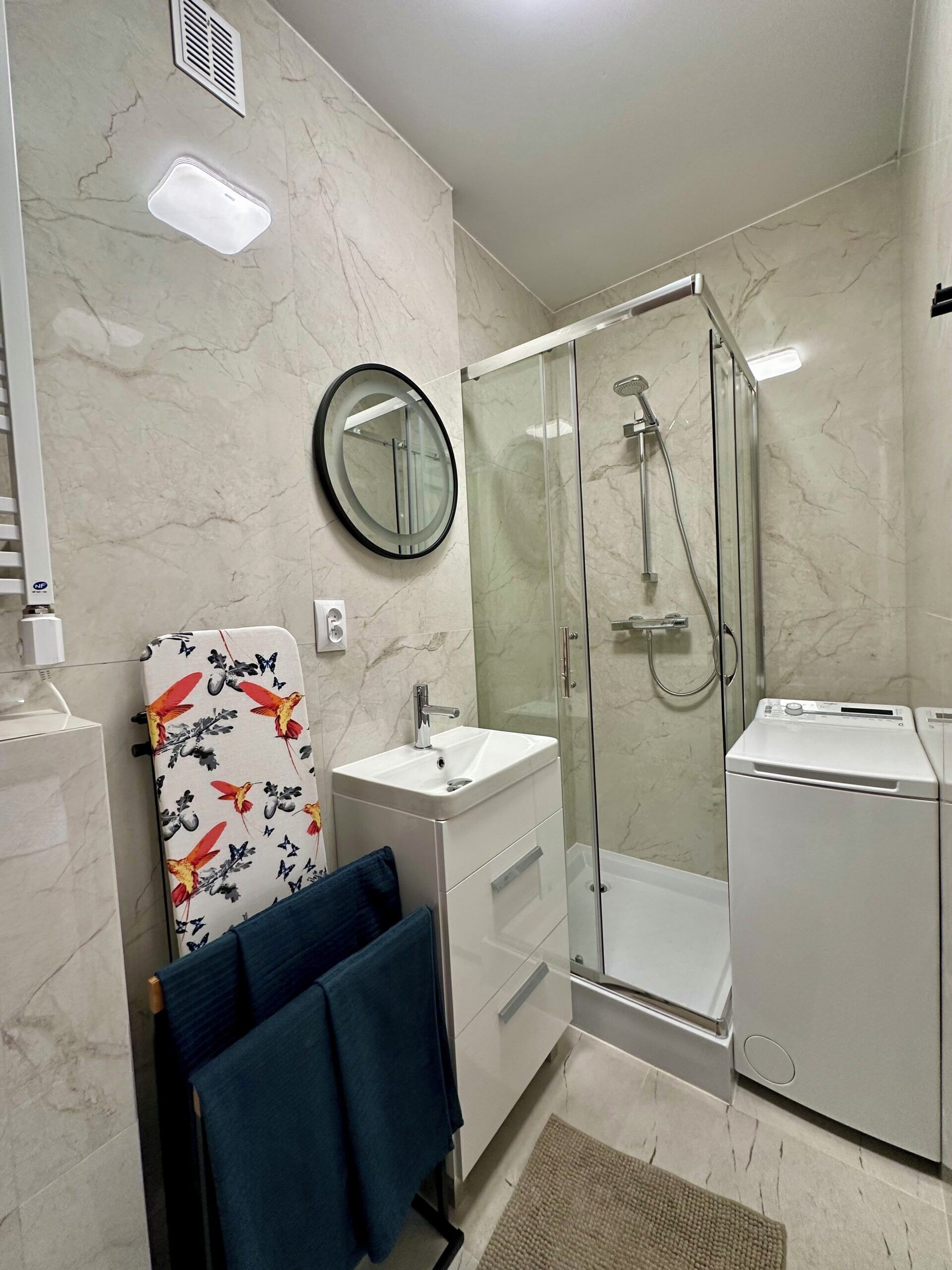 A modern bathroom with a glass shower enclosure, a round mirror, a white sink, and a compact washing machine.