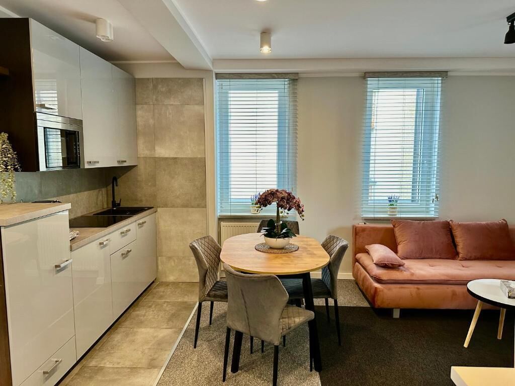 Una cocina moderna con armarios blancos, horno empotrado y un comedor con mesa y sillas redondas. Hay un sofá y una ventana con persianas en la sala.