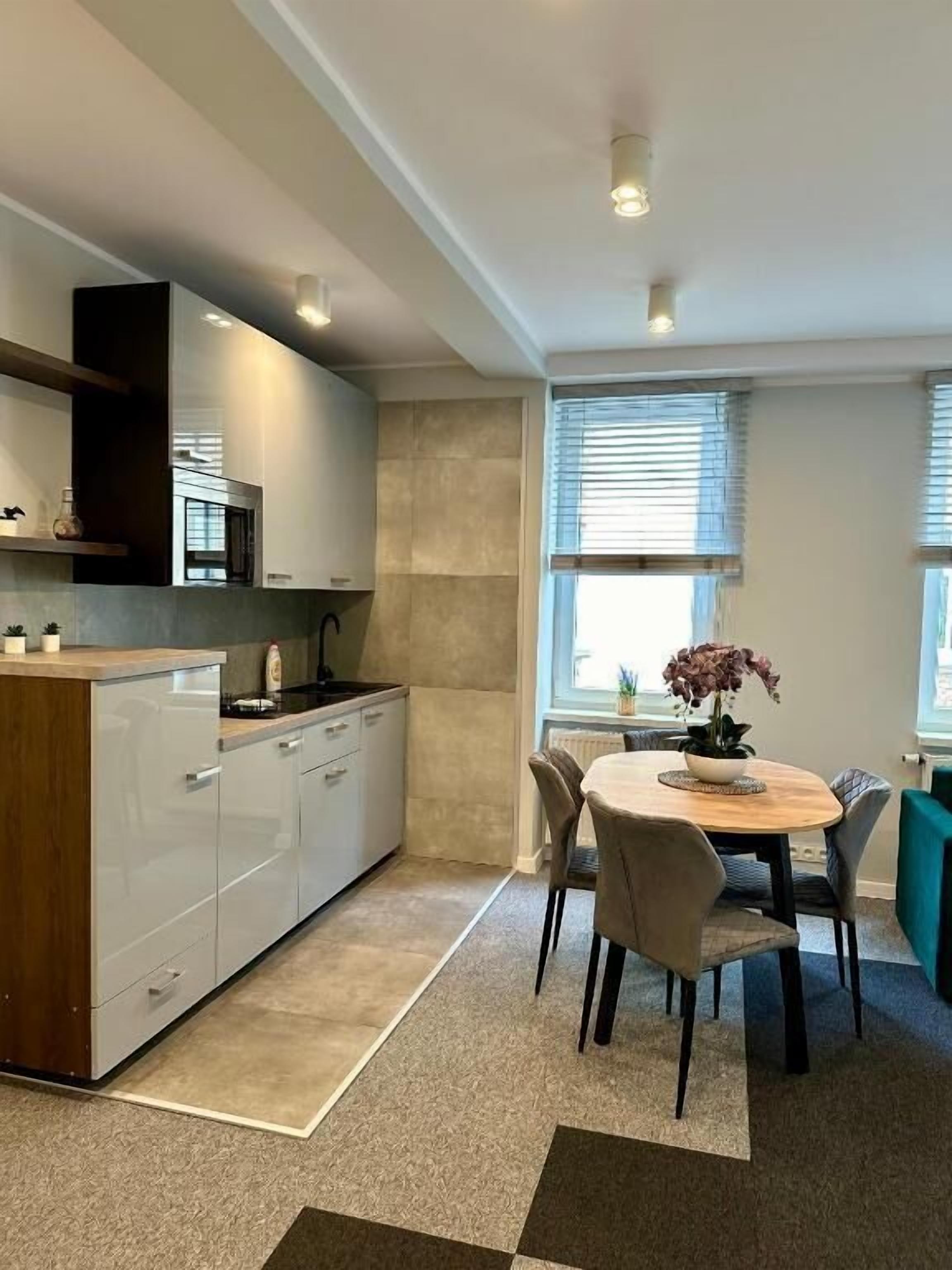 A modern kitchen with white cabinets, a wooden countertop, and a built-in oven. There is a dining area with a round table and chairs, and a window with blinds.
