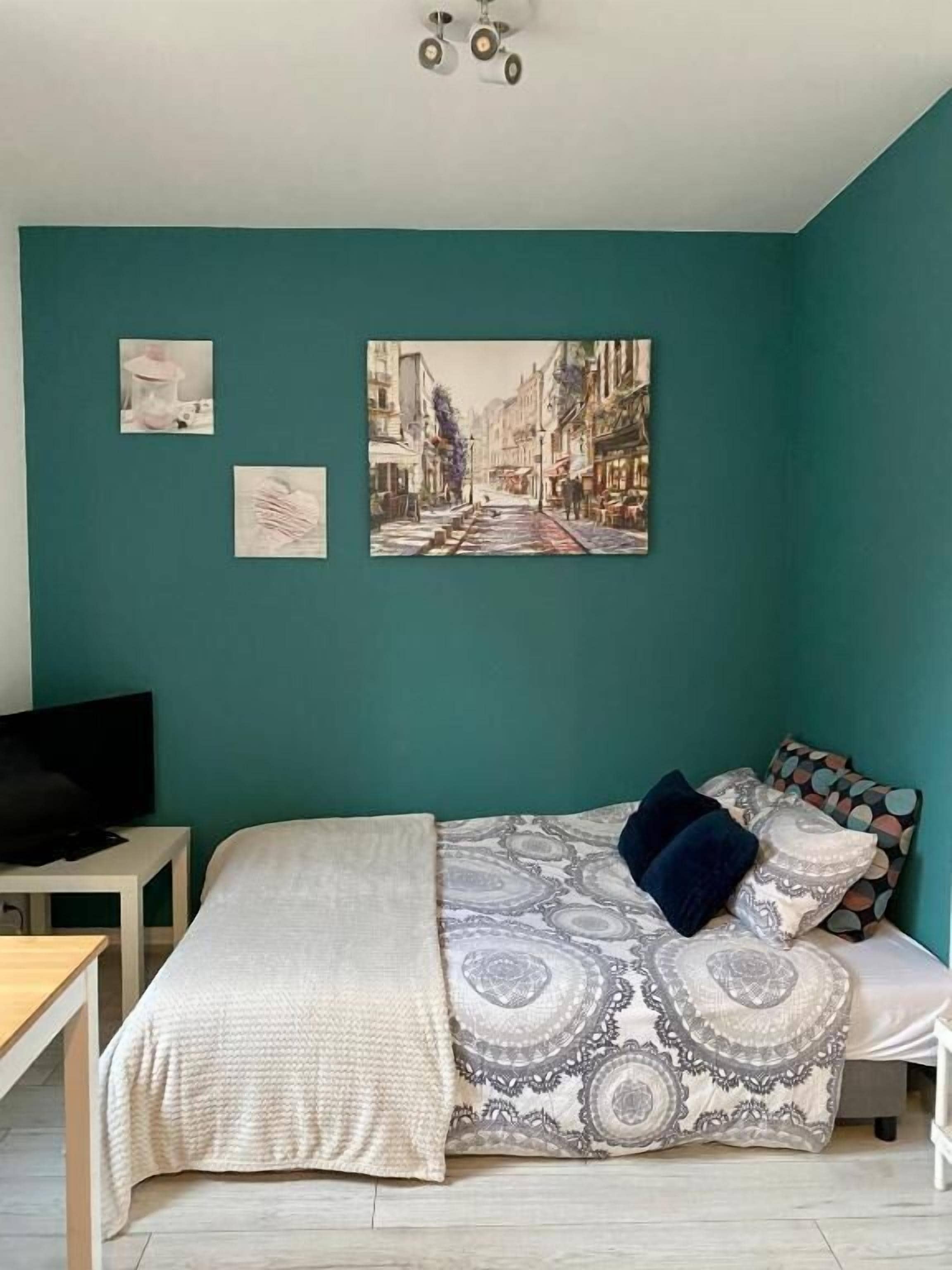 Een slaapkamer met blauwgroene muren, een bed met beddengoed met een patroon, een televisie op een bijzettafeltje en een stadsgezicht schilderij boven het bed.