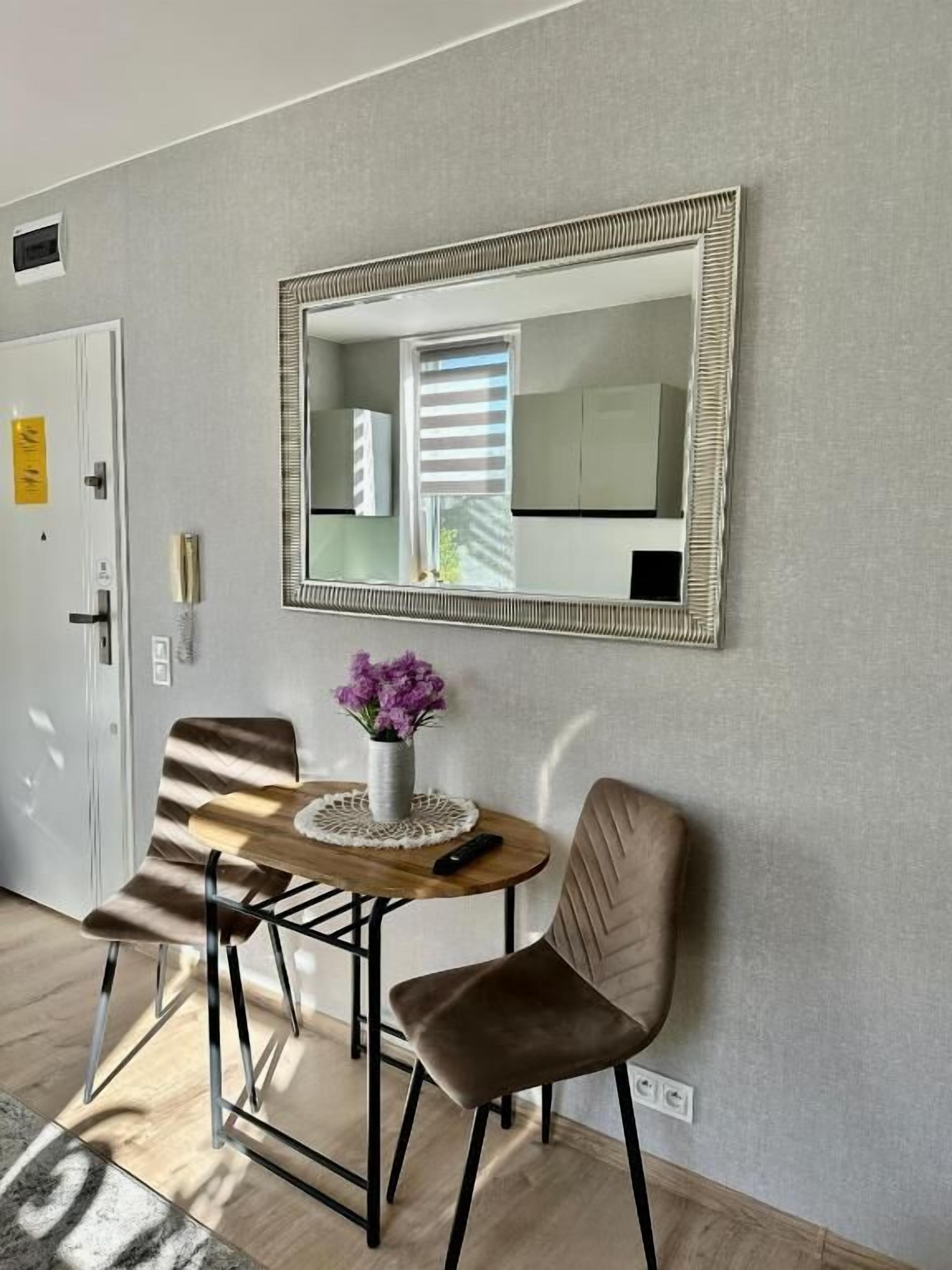A room with a wooden table, two chairs, a vase with purple flowers, and a large mirror on the wall.