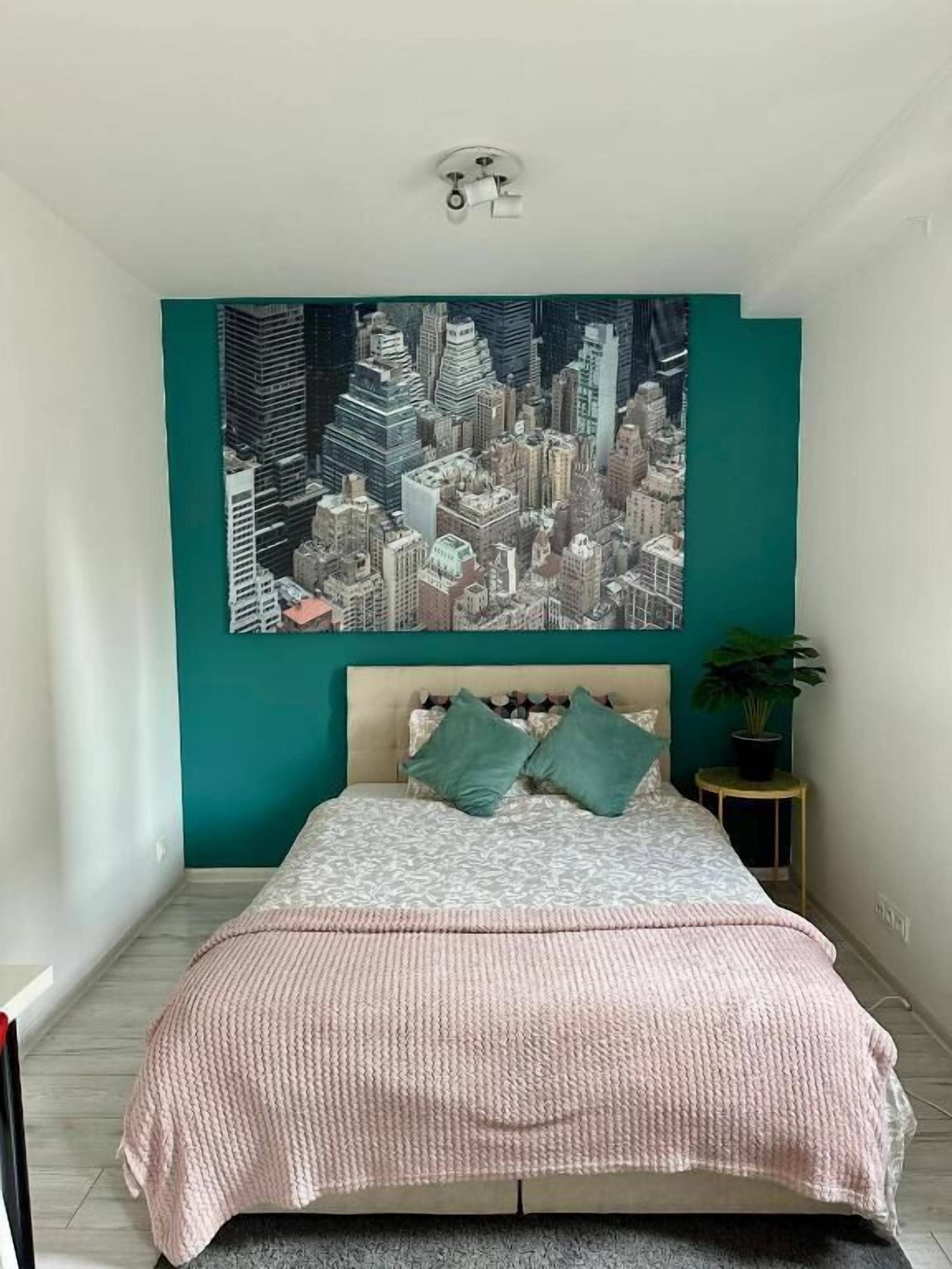 Une chambre avec un mur d’accent de couleur turquoise, une œuvre d’art représentant un paysage urbain, un lit avec une literie à motifs et une petite table ronde sur laquelle est posée une plante.