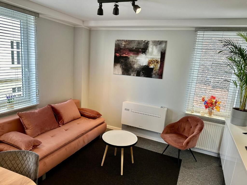 Un salon moderne avec un canapé, une table basse ronde et un fauteuil. On y trouve également un radiateur et une fenêtre avec des stores.