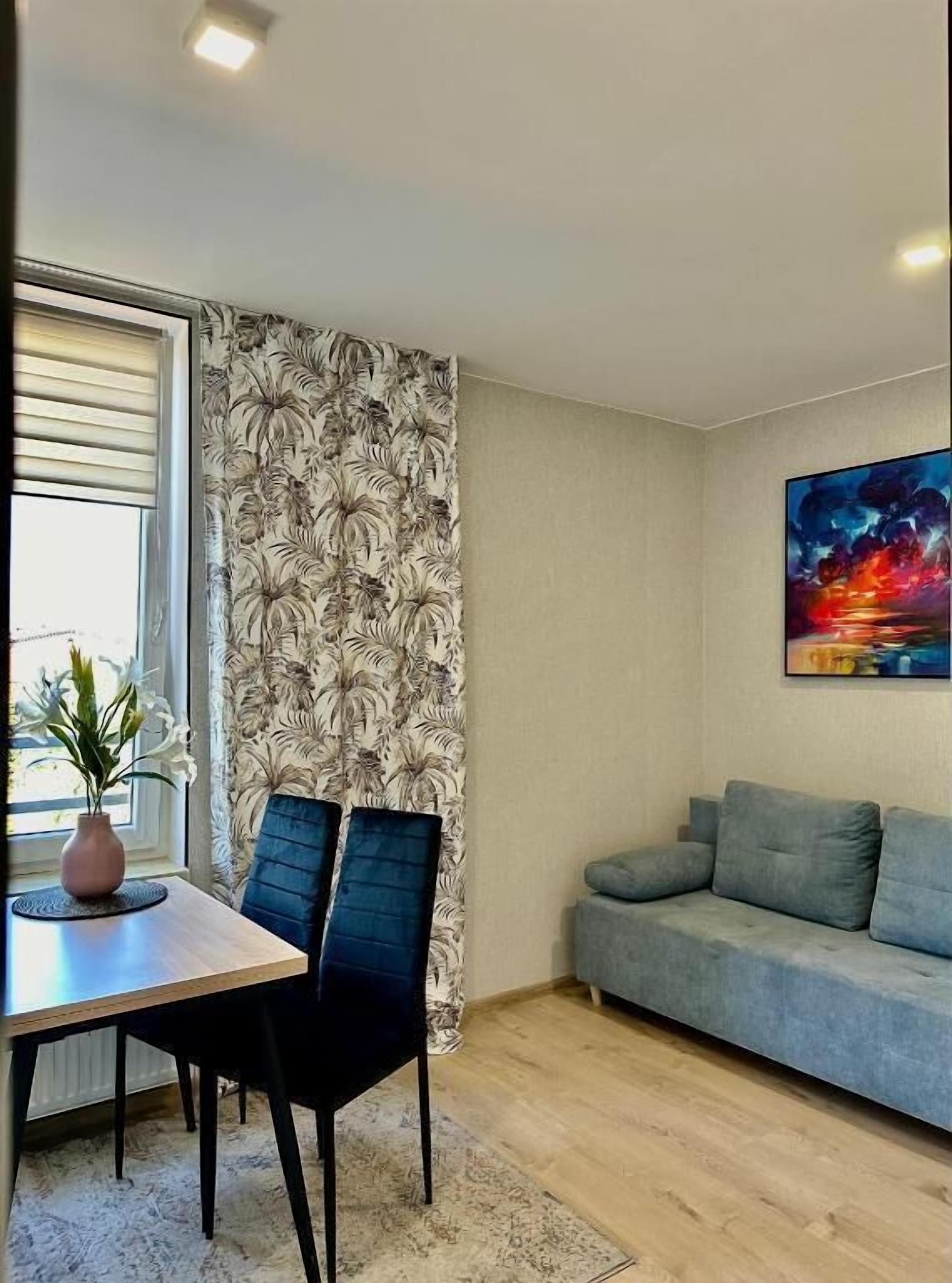Een moderne woonkamer met een eethoek, met een bank, een tafel met stoelen, een raam met zonwering en een schilderij aan de muur.