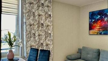 Een moderne woonkamer met een eethoek, met een bank, een tafel met stoelen, een raam met zonwering en een schilderij aan de muur.