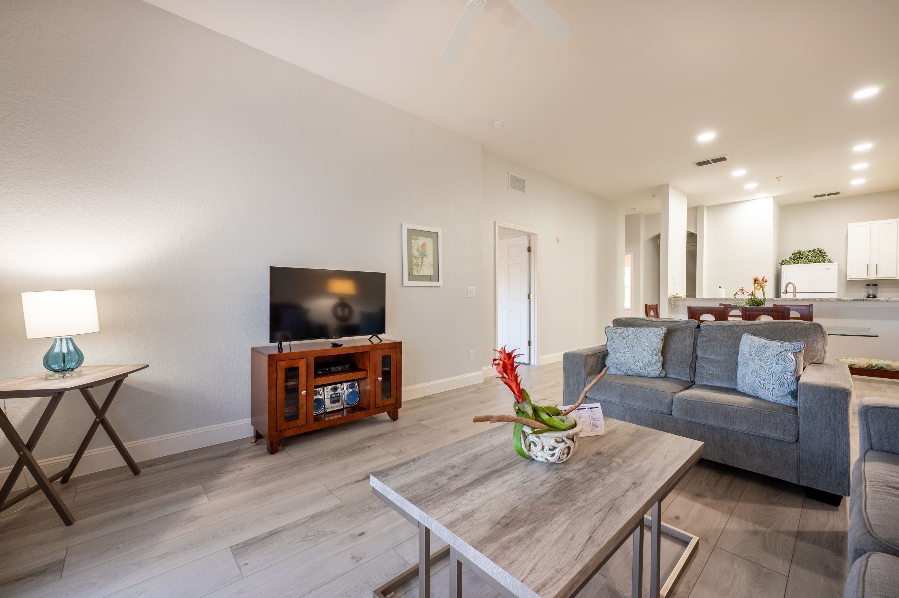 A modern living room with a flat-screen TV, a wooden coffee table, a grey sofa, and a small side table with a lamp.