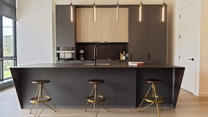 A modern kitchen with a central island, three stools, pendant lights, and a view of the outdoors.