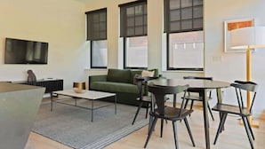 A modern living room with a green sofa, a round dining table with chairs, a flat-screen TV mounted on the wall, and large windows with blinds.