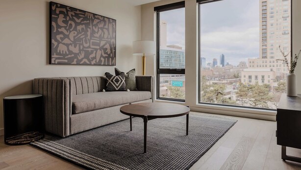 A modern living room with a sofa, a coffee table, a large window with a city view, and a decorative wall piece.