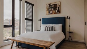 A modern bedroom with a large bed, a wooden bench, two bedside tables, and a city view through large windows.