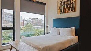 Une chambre d’hôtel moderne avec un grand lit, un banc en bois et une vue sur la ville à travers de grandes fenêtres.