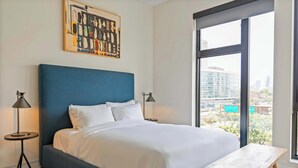 A modern bedroom with a large bed, two bedside lamps, a wooden bench, and a city view through large windows.