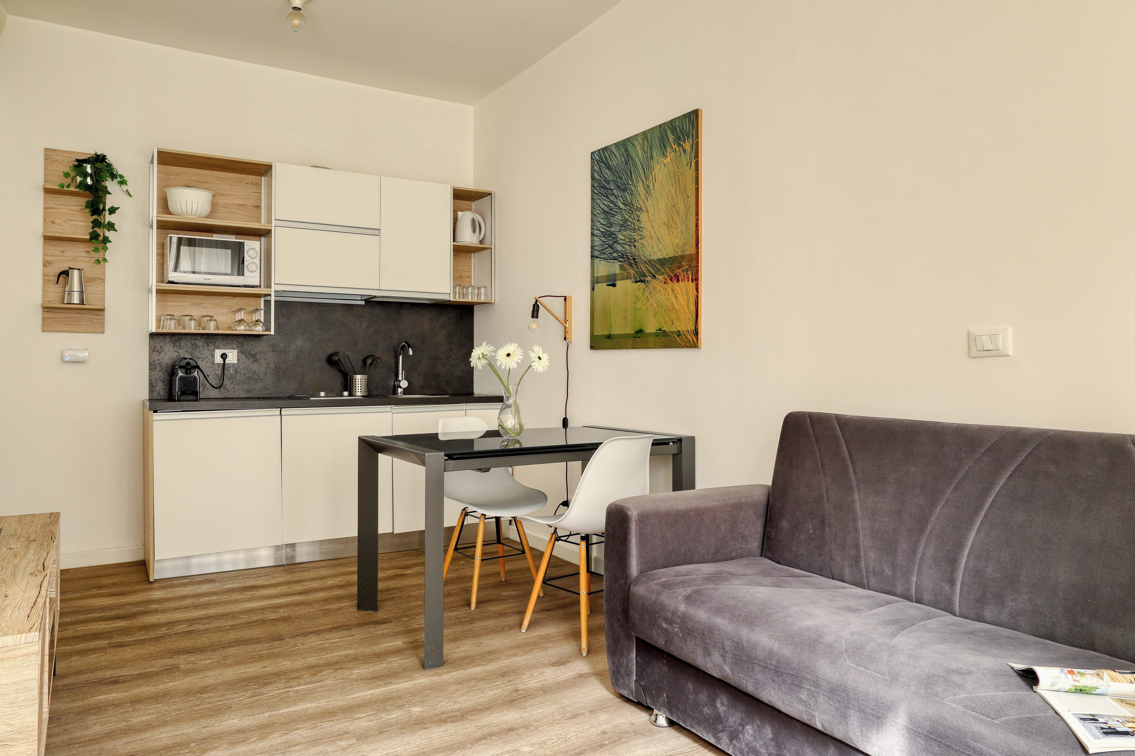 A modern kitchen with white cabinets, a microwave, and a coffee maker. There is a dining table with chairs and a gray sofa in the living area.