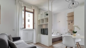 A modern living room with a grey sofa, a dining area with a white table and chairs, and a built-in TV shelf. - Delightful studios near Parco Tre Torri (Milan)