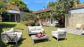 An outdoor seating area with a wooden table and two white sofas, a stone wall, and a building with a tiled roof in the background. - Villa Oscar (Les Mesnuls)