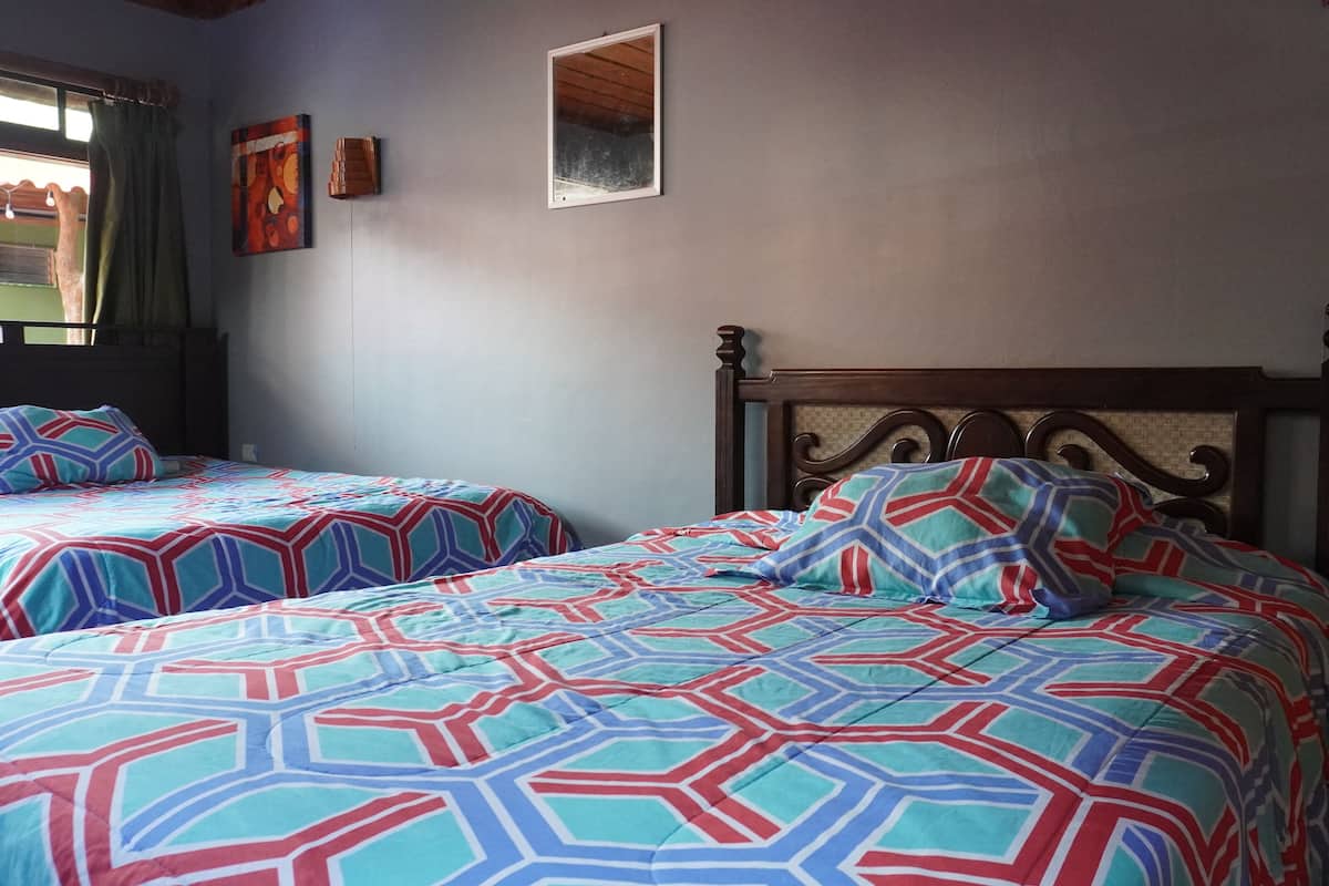 Una habitación con dos camas, cada una con ropa de cama estampada. Hay una ventana con cortinas, un cuadro enmarcado en la pared y un cabecero de madera.