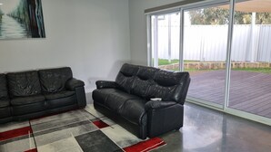 A living room with a black leather sofa, a patterned rug, and a large sliding glass door leading to an outdoor deck. - City View 3 Bedrooms & Work from Home Office, close to airport (Koongamia)