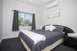 A bedroom with a bed, a window with curtains, an air conditioner, and a painting on the wall.