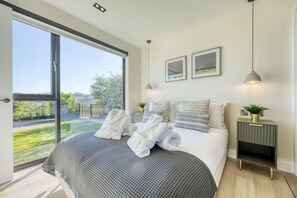 Un dormitorio moderno con un ventanal grande, una cama con toallas enrolladas, mesitas de noche y una planta pequeña.