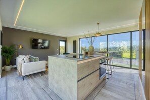 A modern kitchen with a central island, a dining area with a table and chairs, a sofa, and a large window offering a view of the outdoors.