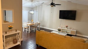 Una sala de estar con un sofá, una mesa de comedor con sillas, un ventilador de techo y un televisor montado en la pared.