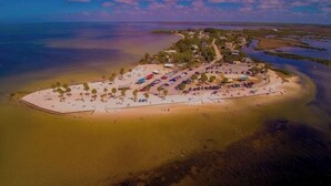 Vista aérea de un complejo turístico frente al mar, con playa, zona de estacionamiento y edificios rodeados de agua.