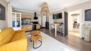 Una sala de estar moderna con un sofá amarillo, un comedor con mesa y sillas, una cocina con armarios de madera y un televisor grande montado en la pared.