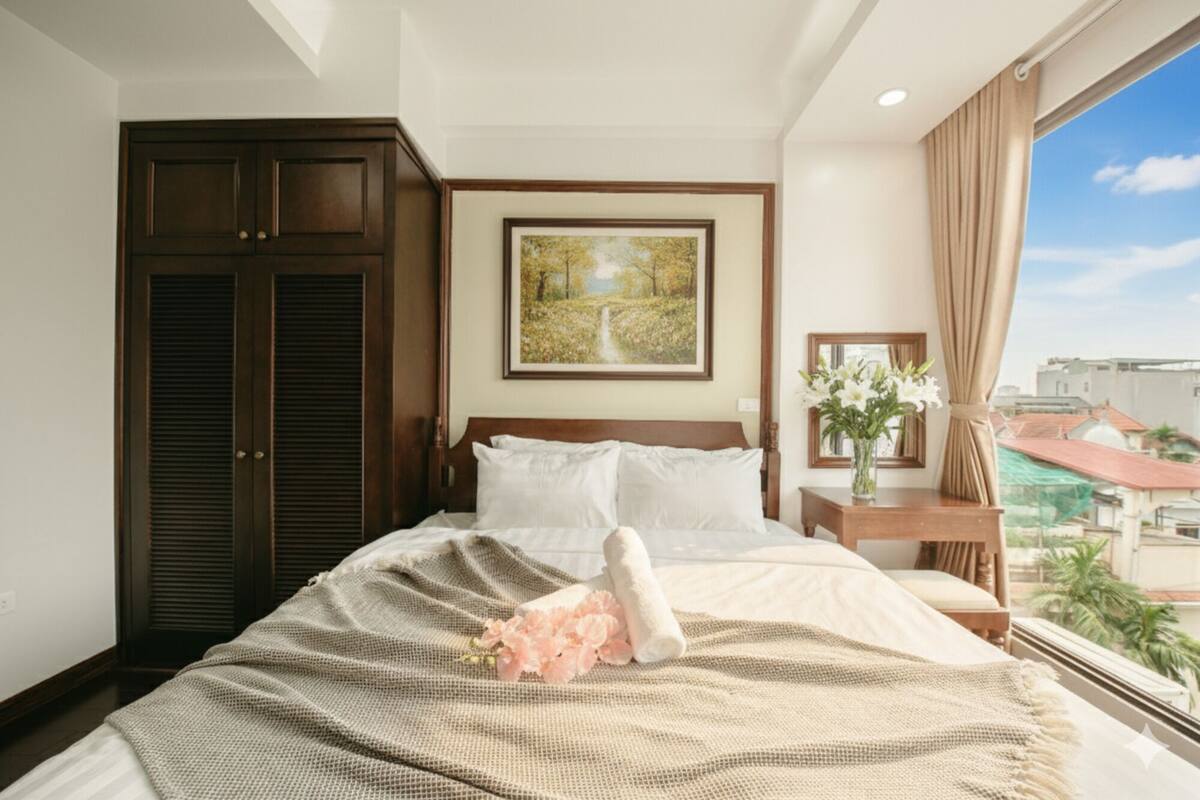 A hotel room with a large bed, a wooden wardrobe, a painting on the wall, a nightstand with a vase of flowers, and a view of buildings through the window.
