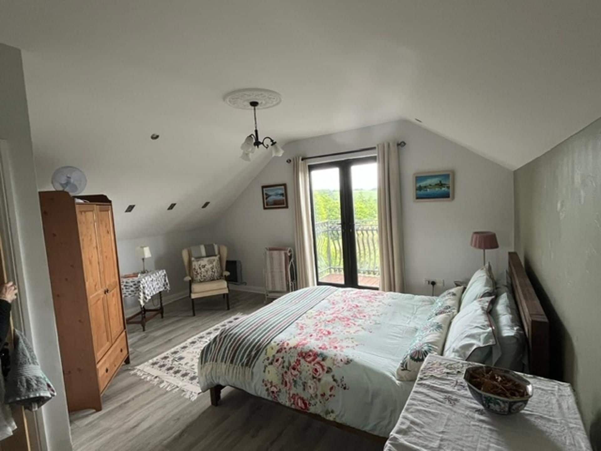 A bedroom with a sloped ceiling, a bed with floral bedding, a wooden wardrobe, a chair, a table with a lamp, and a view through a window.