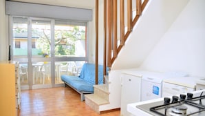 A compact kitchen with a gas stove, a washing machine, and a dining area with plastic chairs. - Cozy Flat With Balcony in Bibione (San Michele al Tagliamento)