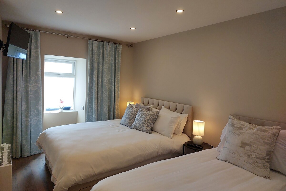 A hotel room with two beds, a TV mounted on the wall, a window with curtains, and a bedside lamp.