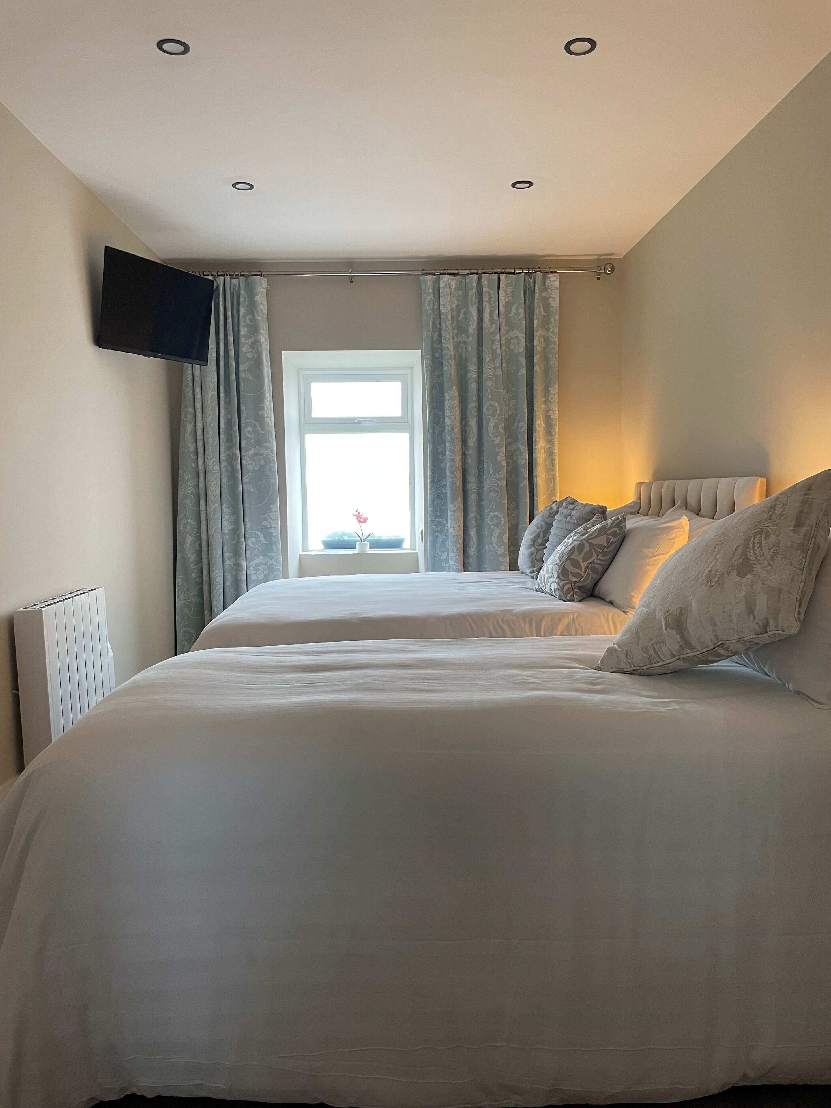 A bedroom with two beds, a TV mounted on the wall, a window with curtains, and a radiator.