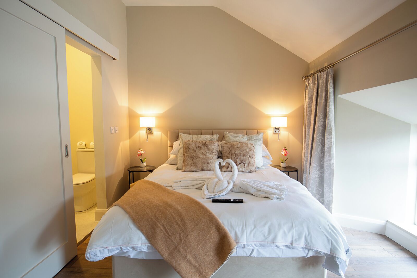 Camera da letto con un letto grande, due comodini, un tavolino e una finestra con tende, oltre a una porta che dà su un bagno.