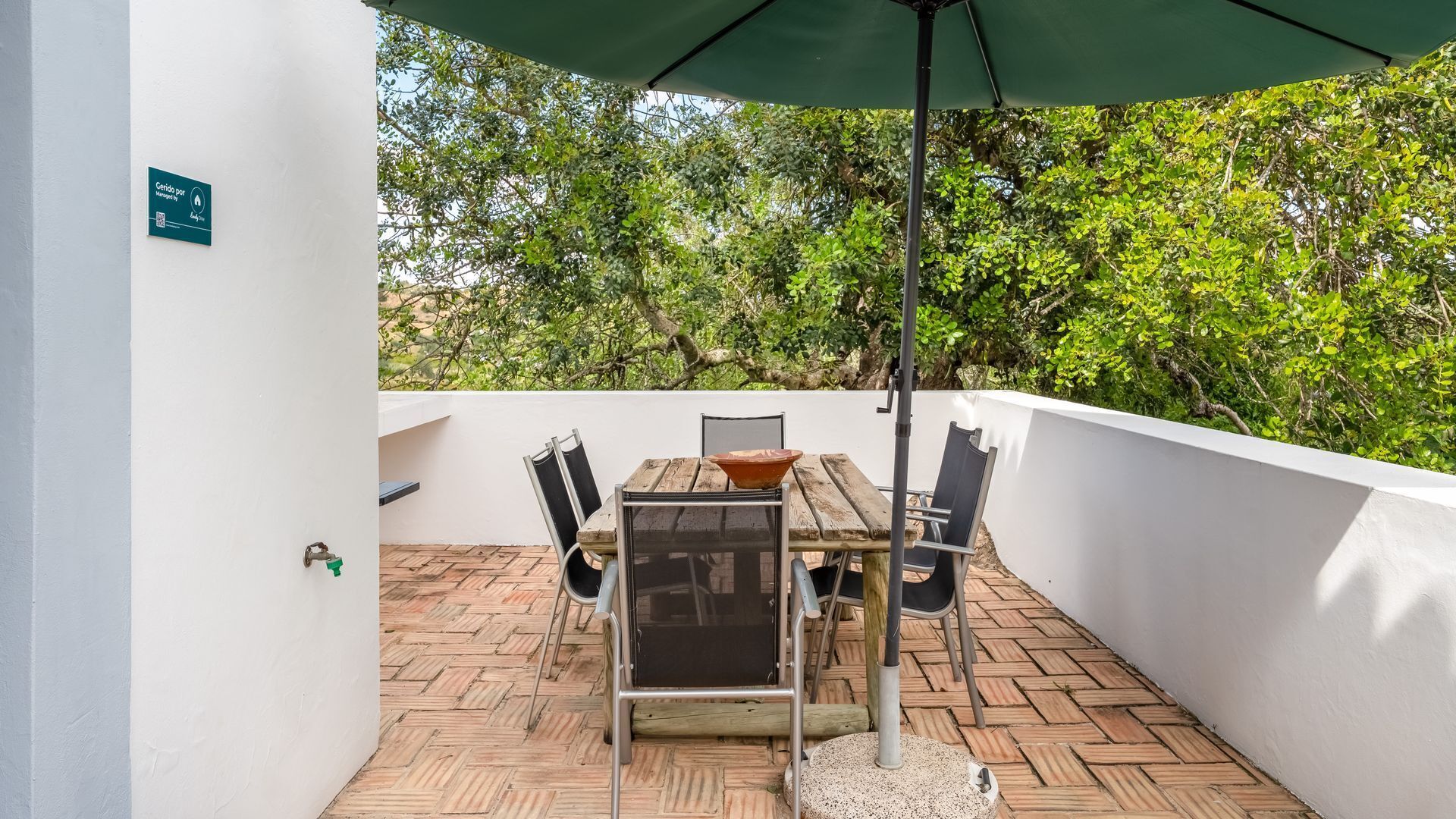 Eine Terrasse mit Holztisch und -stühlen, einem grünen Sonnenschirm und einer weißen Wand mit einem Schild.