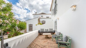 Un patio estrecho con un muro blanco, un muro de piedra, suelo de baldosas, un banco con cojines y una pequeña mesa.
