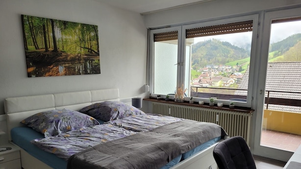 A bedroom with a bed, a window with a view of a village, a radiator, and a small table with a lamp.