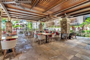 Un'area patio esterna con pavimento in pietra, sedie in vimini e tavoli con cuscini rossi.