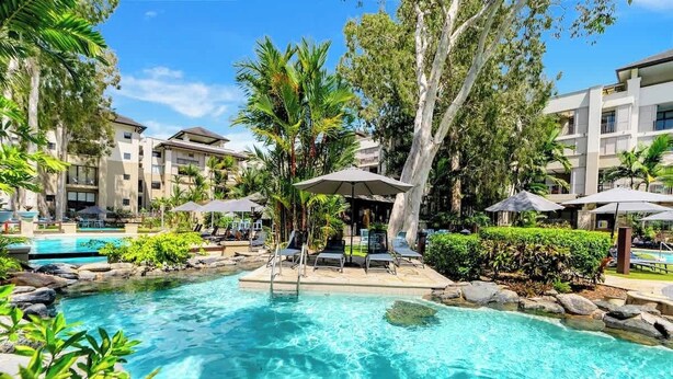 Un espace piscine doté de chaises longues, de parasols et d’une piscine à l’eau d’un bleu limpide, le tout entouré d’arbres et de bâtiments.