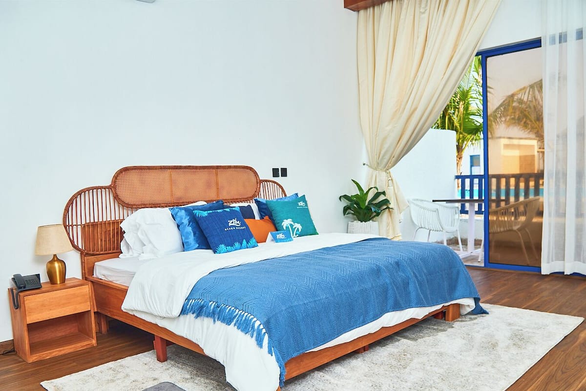 A neatly arranged bedroom with a wooden bed, white bedding, and a blue throw blanket. There is a small bedside table with a lamp and a phone, and a large window with curtains.