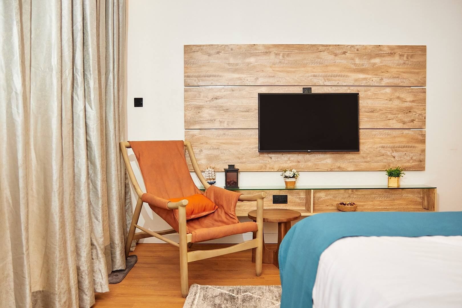 A hotel room with a wooden wall panel, a flat-screen TV, a wooden chair, a small round table, a bed, and a desk with a plant.