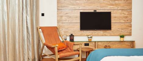 A hotel room with a wooden wall panel, a flat-screen TV, a wooden chair, a small round table, a bed, and a desk with a plant.