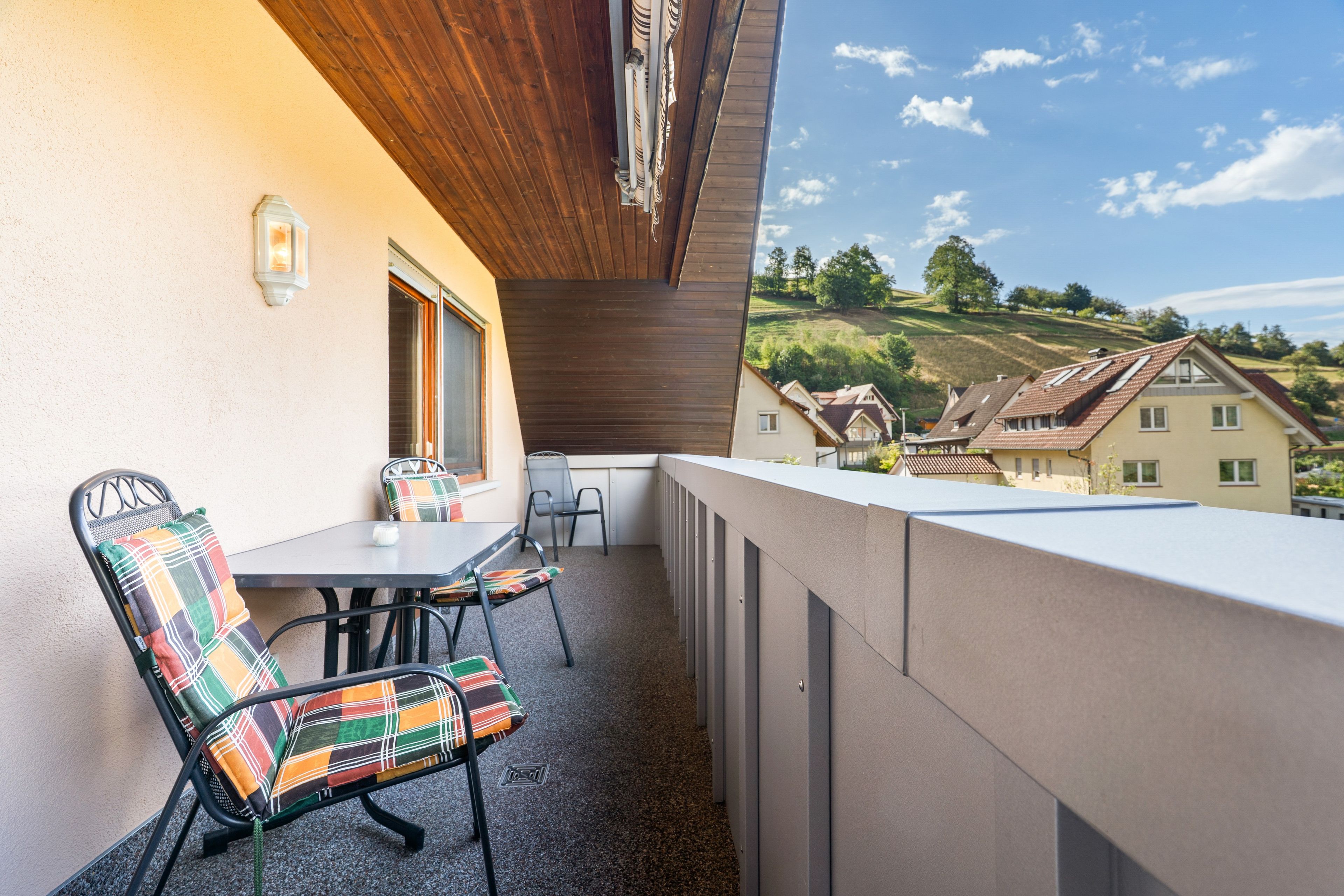 A balcony with a table and chairs, a view of a hillside with houses, and a wooden ceiling.