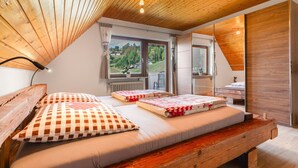Een slaapkamer met een houten plafond, twee bedden met geruit beddengoed, een raam met uitzicht op bomen en huizen, en een radiator.