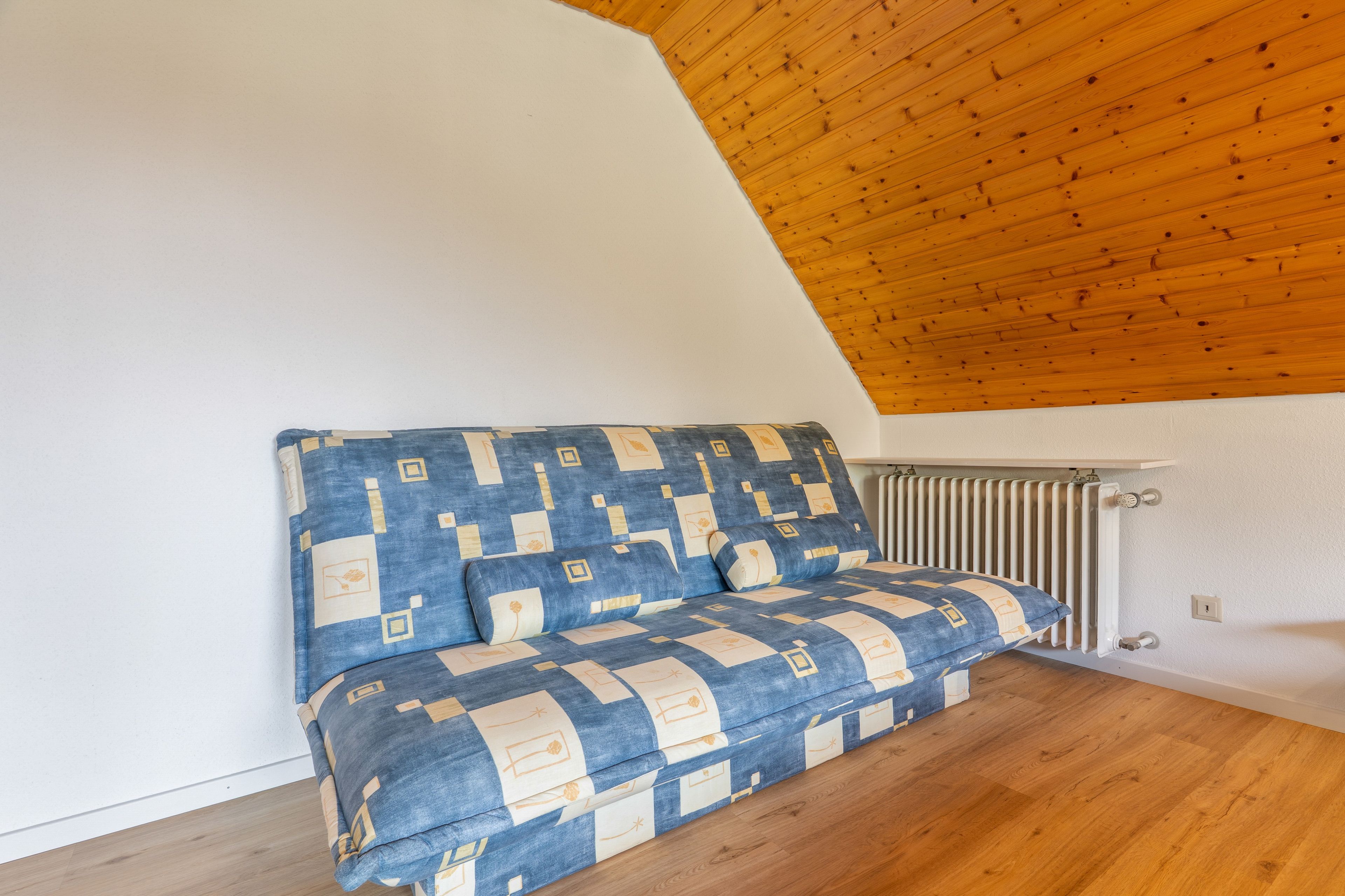 A corner room with a sloped ceiling, a radiator, and a sofa with a geometric patterned cover.
