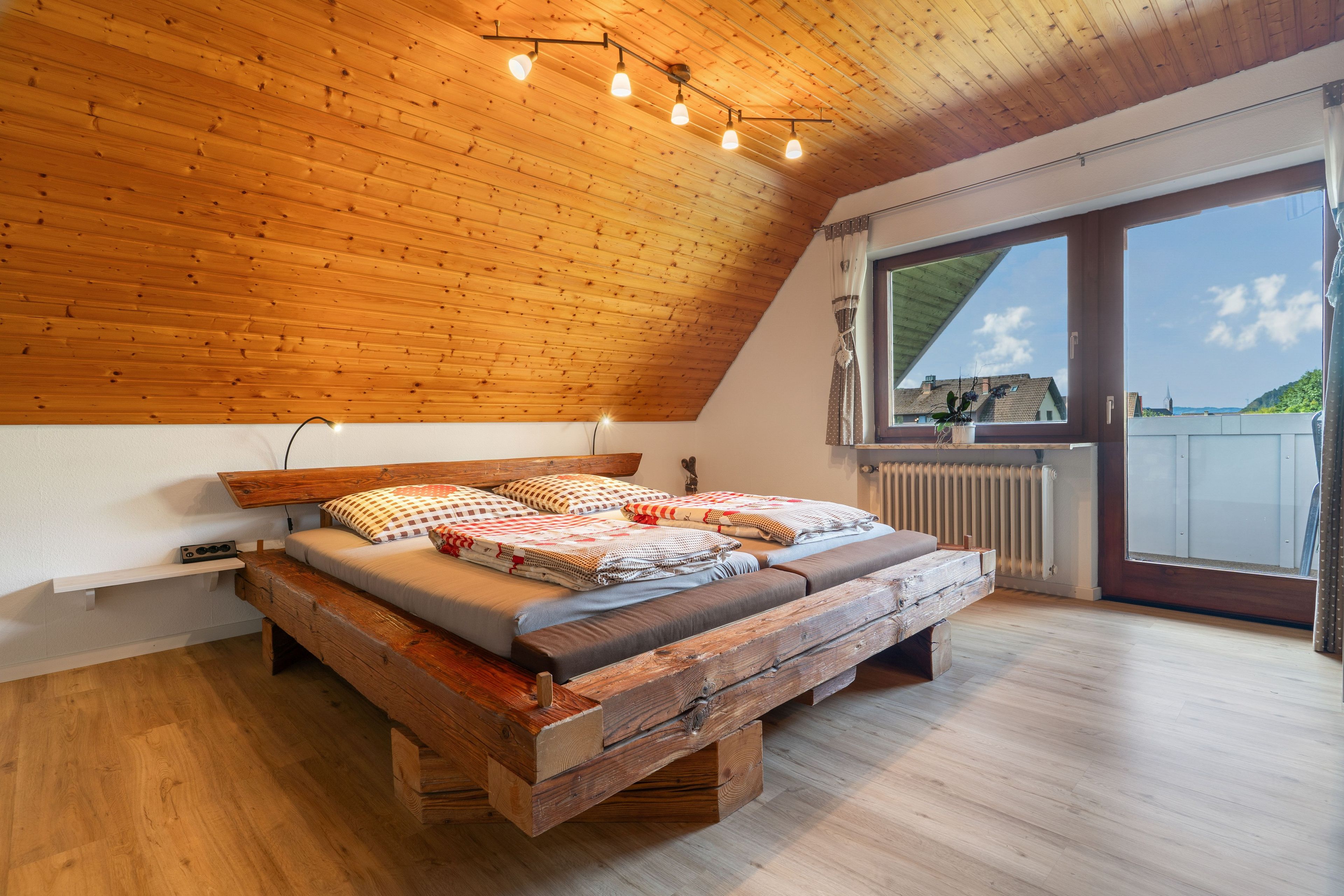 Une chambre de style « loft », avec un plafond en pente, un cadre de lit en bois et une vue sur l’extérieur grâce à une grande fenêtre.