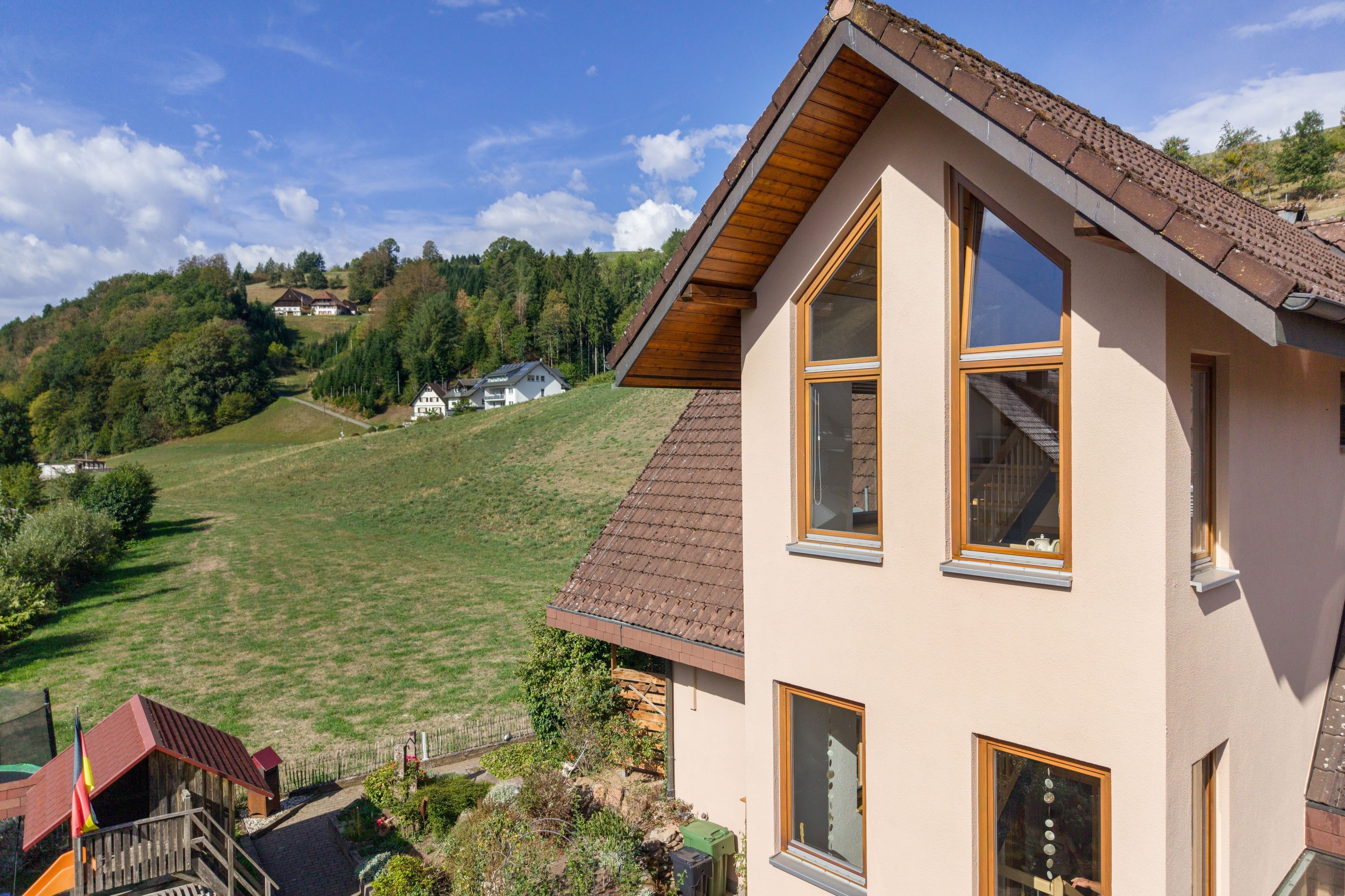 Un edificio a due piani con tetto spiovente, ampie finestre e vista su un pendio verde con alberi e case sullo sfondo.