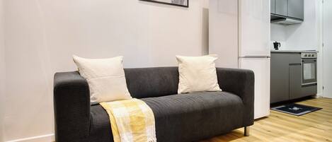 A modern living room with a dark grey sofa, two white cushions, and a yellow and white checkered throw blanket. A kitchen with a white refrigerator, stainless steel oven, and grey cabinets is visible in the background.