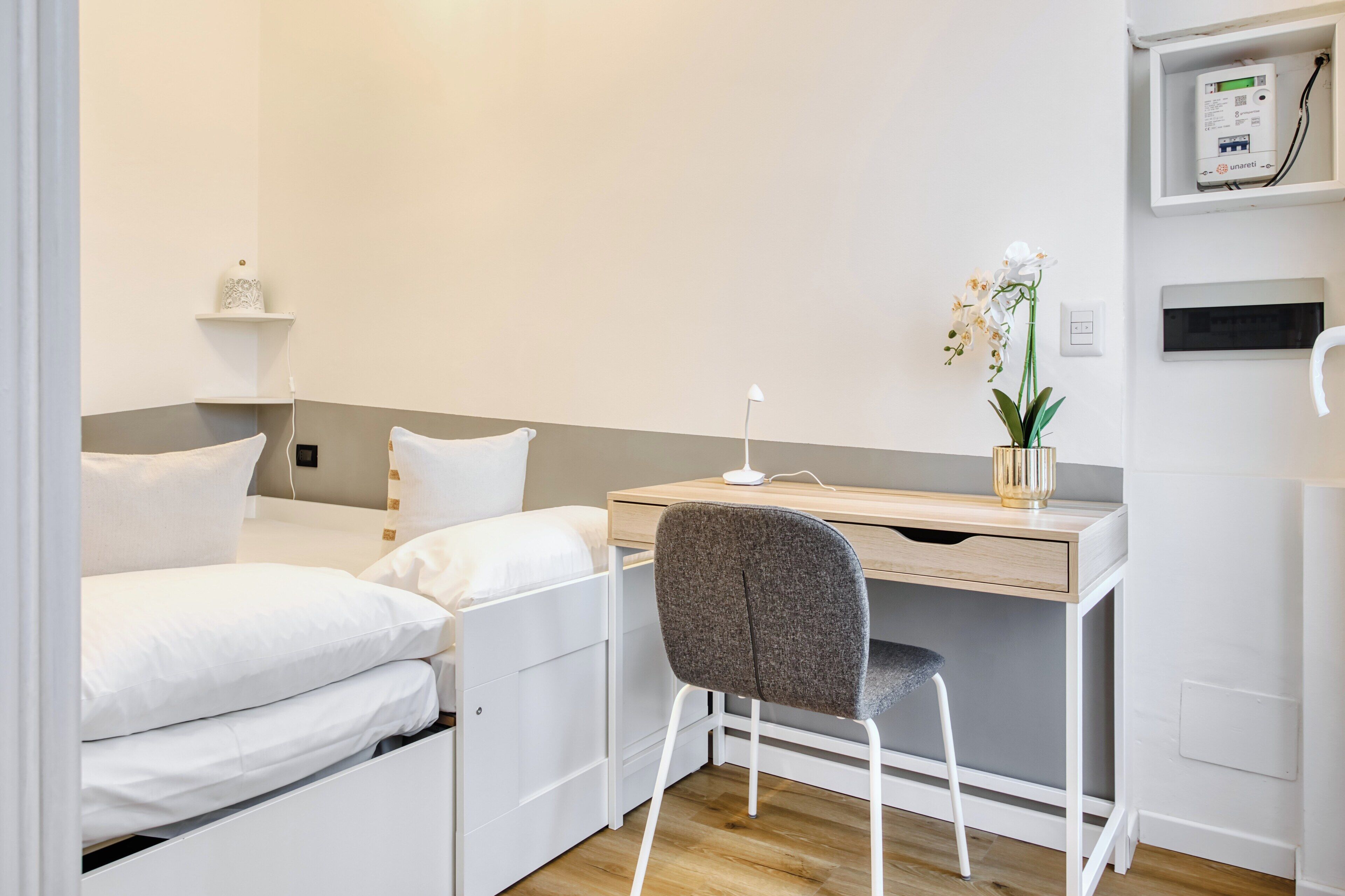 Une chambre à coucher de taille réduite, comprenant un lit, un bureau et une chaise. On trouve une petite étagère sur le bureau, ornée d’un objet décoratif et d’une plante en pot.