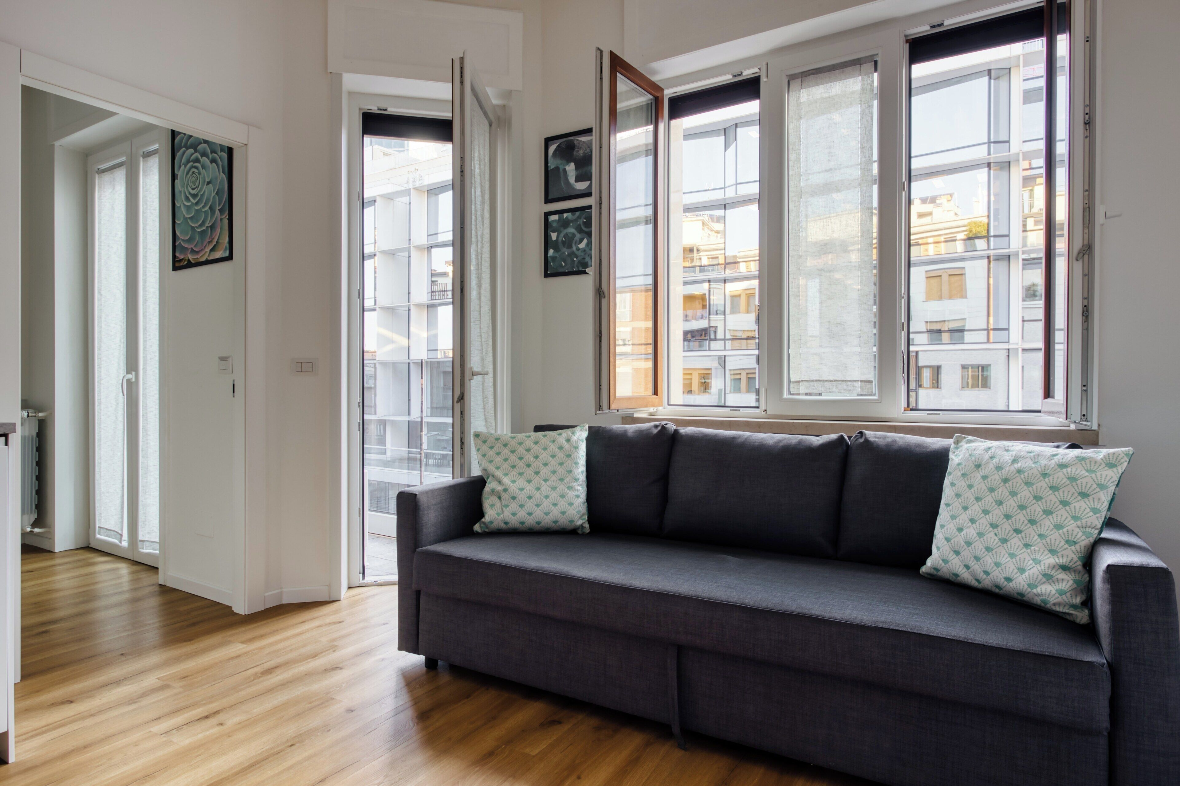 Uma sala de estar moderna com um sofá cinzento, duas almofadas decorativas estampadas e uma grande janela com vista para edifícios.